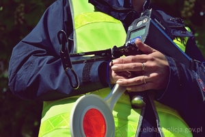 Umundurowny policjant w kamizelce odblaskowej zielonej z policyjnym lizakiem oraz miernikiem prędkości w rękach. Zdjęcie kadrowanie tylko od szyi do pasa. Ilustracja do tekstu.