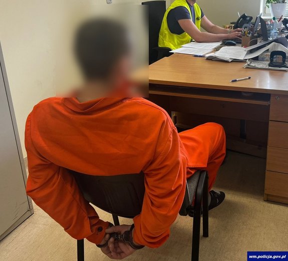 Męzczyzna siedzi tyem na krześle ubrany w pomarańczowy kombinezon. To podejrzany. W tle policjant przy biurku przyjmuje wyjasnienia od zatrzymanego