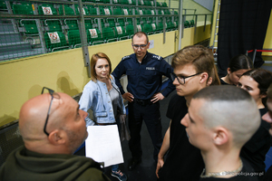 Grupa osób obsługujących interesantów i chętnych do wstąpienia w szeregi Policji. W centralnej części stoi umundurowany policjant