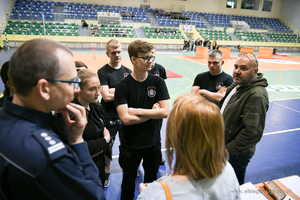 Kobiety i mężczyźni w wieku około 18 lat stojący na hali słuchaja umundurowanego funkcjonariusza.