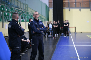 Od lewej strony dwóch umundurowanych policjantów. Mężczyzna po prawej to Komendant Miejski Policji w Elblągu - inspektor Robert Muraszko. w tle hala sportowa z siedziskami na kibiców