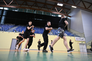 Młodzież szkolna prowadzi rozgrzewkę przed testami sprawnościowymi. Biegają wokół hali sportowej