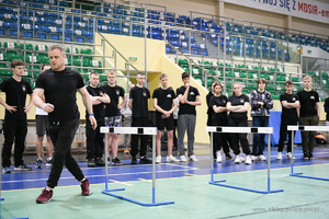 Policyjny instruktor w stroju sportowym tłumaczy grupie kilku osób jak pokonać tor sprawnościowy. W tle są materace i skrzynie drewniane.