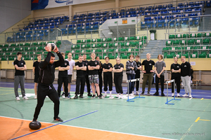 Policyjny instruktor w stroju sportowym pokazuje jak należy rzucać piłką lekarską. W tle są materace i skrzynie drewniane.