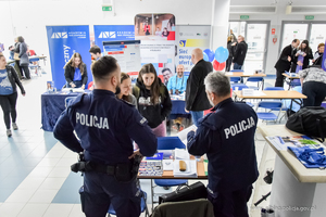 Dwóch umundurowanych policjantów stoi przy stoliku z ulotkami policyjnymi. Rozmawiają z dwiema, młodymi kobietami.