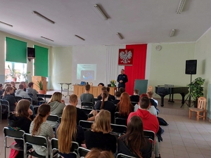Młodzież siedząca w auli szkoły, w tle policjant prowadzący zajęcia.