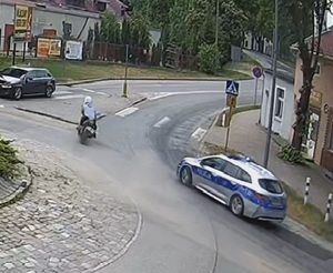 Na zdjęciu widać motocyklistę przejeżdżającego przez rondo a z nim podąża policyjny radiowóz na sygnałach