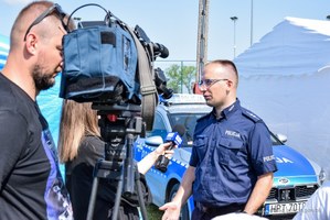 Z prawej strony zdjęcia umundurowany policjant stoi przed kamerą telewizji i udziela wywiadu. Po lewej stronie operator kamery i dziennikarka.