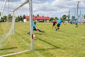 Bramka piłkarska z ujęcia od tyłu. Chłopiec broni strzelonego gola. W tle grupa chłopców pod bramką.