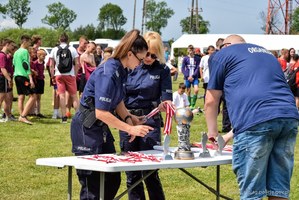 Dwie umundurowane policjantki i mężczyzna nachylają się nad stolikiem sędziowskim, na którym znajdują się medale i puchary dla uczestników zawodów.