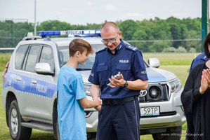 Po lewej stronie kadru chłopiec w stroju sportowym odbiera z rąk umundurowanego policjanta inspektora Roberta Muraszko, Komendanta Miejskiego Policji w Elblągu medal. W tle oznakowany policyjny radiowóz.