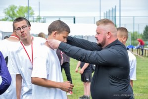 Po lewej stronie kadru chłopiec w stroju sportowym odbiera z rąk mężczyzny w czarnej koszuli medal. Ten mężczyzna to Mateusz Zabłocki, asystent podsekretarza stanu w Ministerstwie Aktywów Państwowych Andrzeja Śliwki.