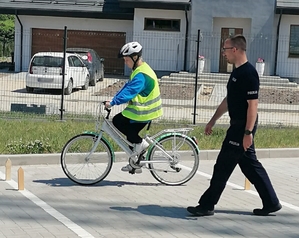 Umundurowany policjant z prawej strony kadru idzie za dzieckie, jadącym rowerem  między pachołkami rowerowymi.