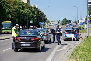 Asfaltowa ulica, dwa ciemne samochody kontrtolowane przez funkcjonariuszy, obok umundurowani policjanci i straznik miejski.