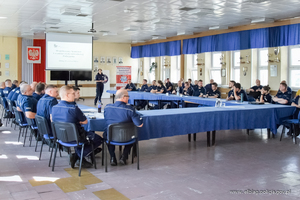 Grupa kilkudziesięciu umundurowanych policjantów siedzi na krzesłach przy stołach. Uczestniczą w wykładzie. W tle stoi umundurowana polijantka i opisuje prezentację multimedialną.
