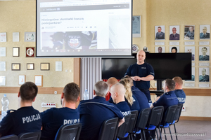 Grupa kilkudziesięciu umundurowanych policjantów siedzi na krzesłach przy stołach. Uczestniczą w wykładzie. W tle stoi umundurowany polijant i opisuje prezentację multimedialną.