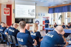 Grupa kilkudziesięciu umundurowanych policjantów siedzi na krzesłach przy stołach. Uczestniczą w wykładzie. W tle stoi umundurowana polijantka i opisuje prezentację multimedialną.
