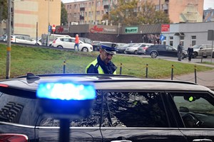 Umundurowany policjant ruchu drogowego kontroluje dokumenty kierującego i bada jego stan trzeźwości. Stoi przy czarny samochodzie typu kombi.