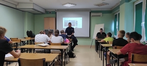Grupa dzieci siedzi w ławkach w sali szkoleniowej. W tle umundurowani policjanci ruchu drogowego.
