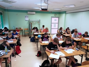 Grupa dzieci siedzi w ławkach w sali szkoleniowej. W tle umundurowani policjanci ruchu drogowego.