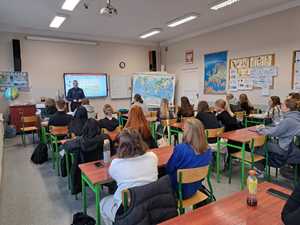 Klasa w szkolen a w niej grupa młodziezy. W tle umundurowany policjant stoi przy tablicy.