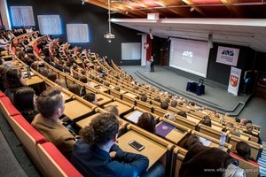 Sala wykładowa dla studentów widziana z góry. Na dole, na scenie umundurowany policjant stoi przy mównicy, obok wyświetlona prezentacja multimedialna.