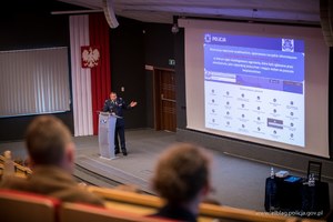 Sala wykładowa dla studentów widziana z góry. Na dole, na scenie umundurowany policjant stoi przy mównicy, obok wyświetlona prezentacja multimedialna.