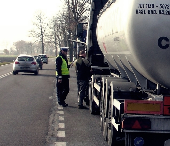 Policjant w mundurze stoi przed zatrzymaną cieżarówką z cysterną paliwa. Kontroluje kierowcy dokumenty oraz stan techniczny pojazdu.