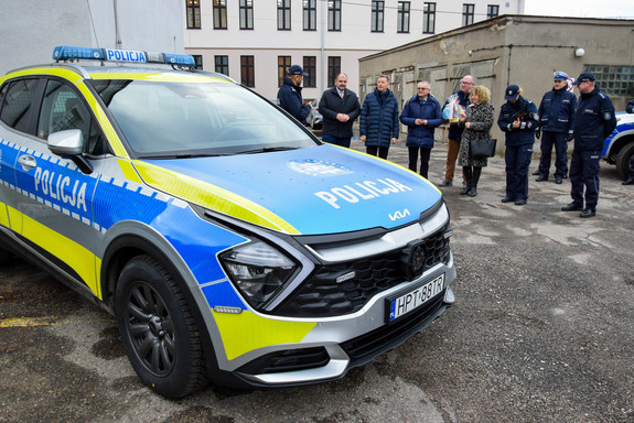 Oznakowany policyjny radiowóz. W tle kilka osób mężczyzn i kobiet