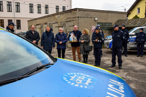 Oznakowany policyjny radiowóz. W tle kilka osób mężczyzn i kobiet