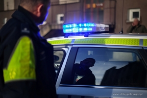 Umundurowany policjant stoi przy oznakowanym radiowozie. Zdjęcie stanowi ilustrację do tekstu.